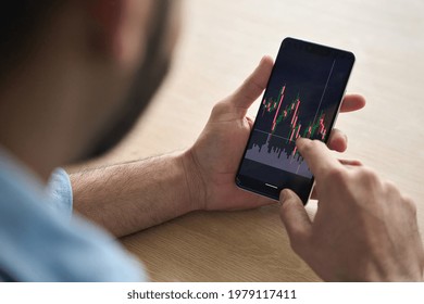 Over shoulder close up view of male analyst broker's hands holding touch screen device smartphone using tech ecommerce application of finance stock markets with graphs and numbers. - Powered by Shutterstock