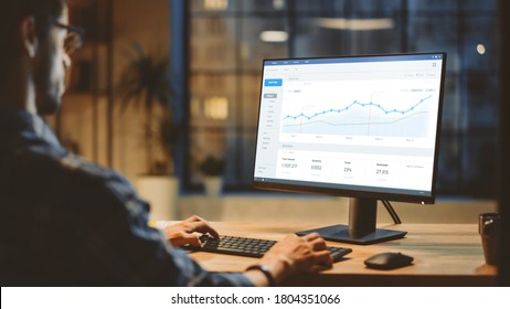 Over The Shoulder Confident Young Business Man Sitting At His Desk Using Desktop Computer With Statistics, Various Data, Charts And Graphs. Evening In Stylish Office Studio With City Window View