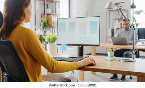 Over The Shoulder: Female Mobile Software Developer Sitting At Her Desk Using Desktop Computer With Screen Showing Smartphone Application UI / UX. Office With Diverse Team Of Professionals Working.