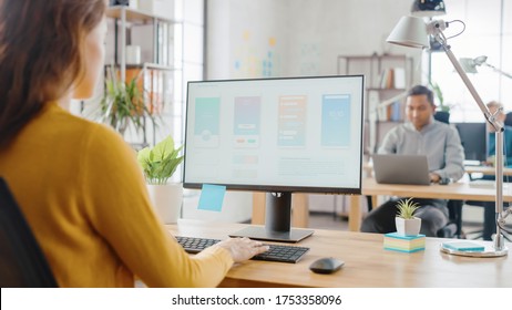 Over The Shoulder: Female Mobile Software Developer Sitting At Her Desk Using Desktop Computer With Screen Showing Smartphone Application UI / UX. Office With Diverse Team Of Professionals Working.