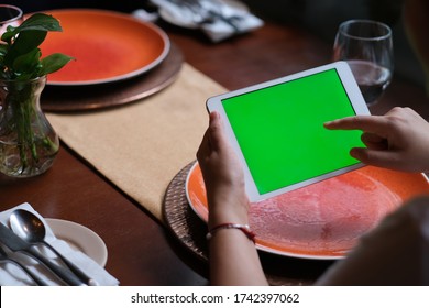 Over Shoulder Of People Tapping Green Screen Tablet In Restaurant. Blurred Plates On Dining Table.