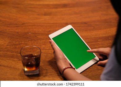 Over Shoulder Of People Tapping Green Screen Tablet. A Glass Of Iced Drink On Wooden Table