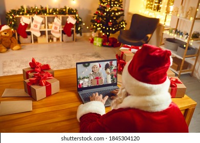 Over Shoulder Santa Claus Sitting At Laptop Computer In His Workshop Having Video Call With Happy Child Holding Xmas Present. Gift-giving, Self-isolation, Virtual Christmas Celebration At Home Concept