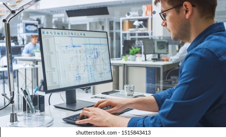 Over The Shoulder Shot Of Engineer Working With CAD Software On Desktop Computer, Screen Shows Technical Drafts And Drawings. In The Background Engineering Facility Specialising On Industrial Design