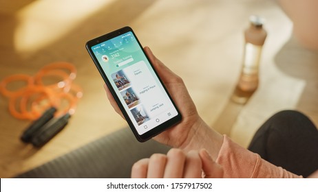 Over Shoulder Shot: Fit Woman Holds Smartphone, Uses Fitness Tracking App, Looking Through Premium Exercise Videos. Mock-up Training Application Software - Powered by Shutterstock