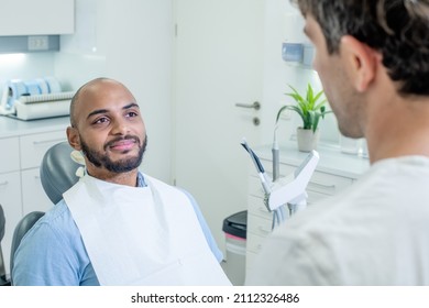 Over The Shoulder Shot Of A Male Dentist Talking Reassuringly To His African American Male Patient, Who Smiling And Looking Relaxed While Sitting In The Dental Chair.