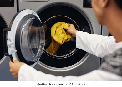 Over shoulder shot of man loading delicate cotton sweaters into washing machine refreshing clothes at coin laundromat, copy space