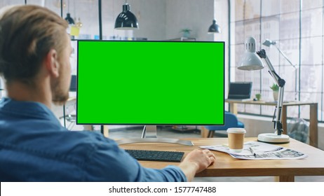 Over The Shoulder Shot: Man Uses Desktop Computer With Green Mock-up Screen While Sitting At His Desk. In The Background Stylish Modern Open Space Creative Business Office