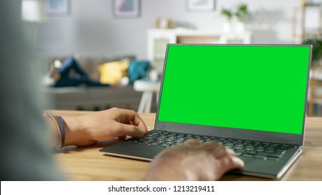 Over the Shoulder Shot of Mans Hands Using Laptop with Green Mock-up Screen. In the Background Cozy Living Room with Woman Relaxing on a Sofa. - Powered by Shutterstock