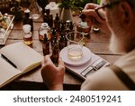 Over shoulder shot of mature perfumer adding extra essential oil while glass flask with perfume sample on kitchen scales according to his notes