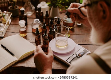 Over shoulder shot of mature perfumer adding extra essential oil while glass flask with perfume sample on kitchen scales according to his notes