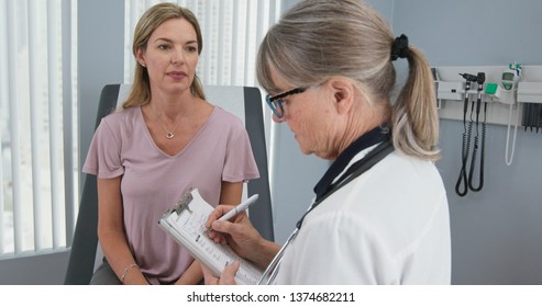 Over The Shoulder Shot Of Woman Talking To Her Primary Care Doctor In Exam Room. Middle Aged Patient Having Appointment With Female Senior Physician