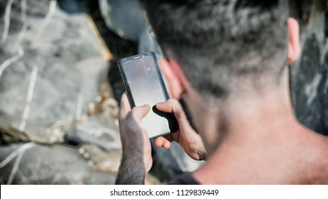 Over The Shoulder Shot Of A Young Man Using Cell Phone To Send Text Message, On A Beach