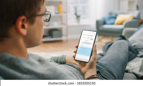 Over the Shoulder Shot of a Young Man at Home Sitting on a Sofa and Using Smartphone for Checking Weather Predictions and Coming Day Plans in Daily Schedule Calendar. Cozy Living Room. - Shutterstock ID 1833205150