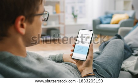 Over the Shoulder Shot of a Young Man at Home Sitting on a Sofa and Using Smartphone for Checking Weather Predictions and Coming Day Plans in Daily Schedule Calendar. Cozy Living Room.