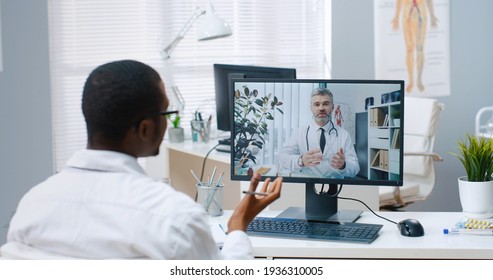 Over Shoulder View Of African American Male Specialist Talks On Online Video Call With Caucasian Middle-aged Doctor On Computer Sitting In Office Experienced Physician Consults Nurse On Web Conference