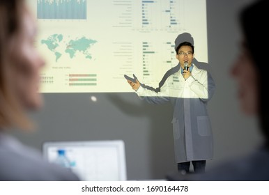 Over Shoulder View Of Asian Speaker In Lab Coat Talking About Benefits Of Project And Pointing At Projection Screen At Conference