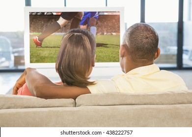 Over Shoulder View Of Casual Couple Watching Tv Against Football Players Tacakling For The Ball On Pitch