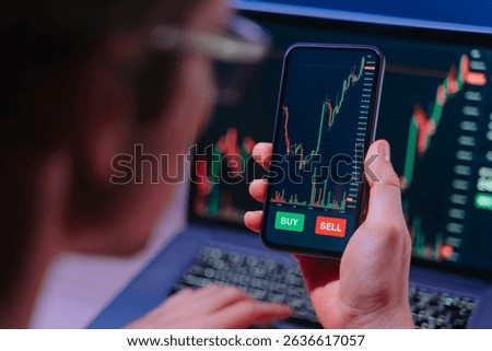 Over shoulder view of crypto trader analyzing live crypto and stock charts on smartphone with trading platform app, displayed on laptop and phone screen in dark neon lit room.