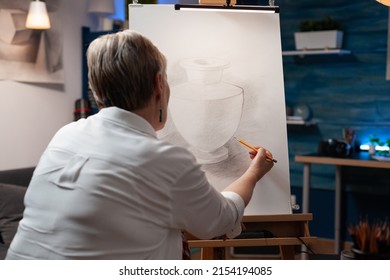 Over Shoulder View Of Elderly Artist Drawing Still Life Using Sharp Pencil On Paper Sketching Vase Model In Home Art Studio. Portrait Of Older Woman Sketching Original Masterpiece In Workshop.