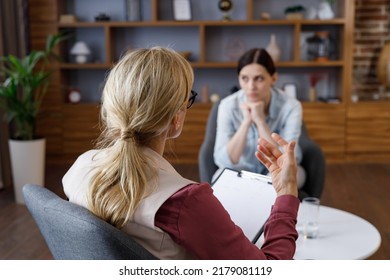 Over shoulder view of female psychologist sitting in armchair, talking with upset woman patient. Psychologist taking notes on clipboard. Psychology, mental therapy, mental health, therapy session - Powered by Shutterstock