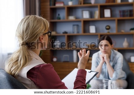 Over shoulder view of female psychologist sitting in armchair, talking with upset woman patient. Psychologist taking notes on clipboard. Psychology, mental therapy, mental health, therapy session