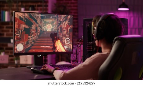 Over Shoulder View Of Gamer Girl Using Pc Gaming Setup Relaxing Playing Multiplayer Online Action Video Game Talking Using Headset. Woman Streaming First Person Shooter While Explaining Gameplay.