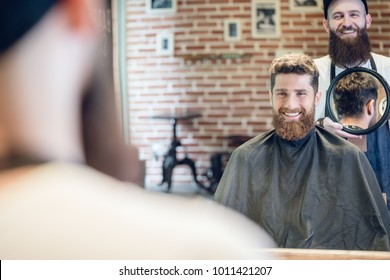 Over the shoulder view of a handsome young man smiling, while looking at his new trendy haircut in the mirror held by his experienced barber in a cool hair salon - Powered by Shutterstock