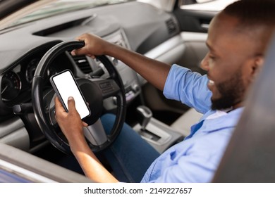 Over the shoulder view of happy black male driver holding smartphone with white blank screen, using mobile app for navigation, mock up. Closeup of cell phone with empty display, copy space template