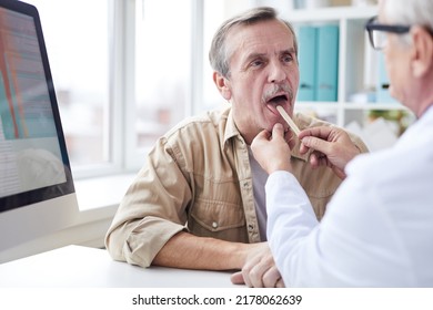 Over Shoulder View Of Male Doctor Sitting At Table And Using Tongue Depressor While Looking At Throat Of Senior Patient