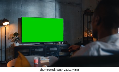 Over The Shoulder View Of Male Watching TV With Mock Up Green Screen Chromakey Display While Sitting On Sofa With Remote Controller In Hands. Man Chilling At Home In The Evening.