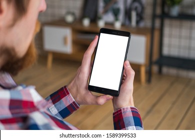 Over shoulder view: man hands holding black smartphone with white blank screen in home interior. Mock up, copyspace, leisure time, template, entertainment and technology concept