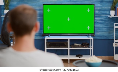 Over Shoulder View Of Man Watching Movie On Tv With Green Screen Relaxing With Bowl Of Popcorn Sitting On Couch. Back View Of Person Relaxing On Sofa In Front Of Television Mockup With Chroma Key