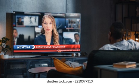 Over The Shoulder View Of Man Watching TV News, Sitting On Couch In Stylish Loft Living Room. TV Presenter Telling Breaking News About Construction Industry. Home With Cozy Interior.