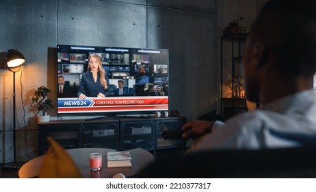 Over The Shoulder View Of Man Watching TV News, Sitting On Couch In Stylish Loft Living Room. TV Presenter Telling Breaking News About Construction Industry. Home With Cozy Interior Concept.