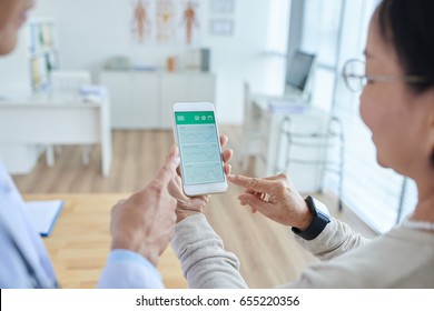 Over Shoulder View Of Middle-aged Physician Showing His Female Patient How To Use Healthcare Mobile App, Interior Of Doctors Office On Background