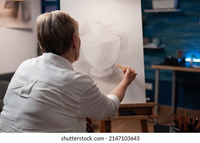 Over shoulder view of older woman drawing orginal masterpiece using sharp pencil on paper sketching vase model in home art studio. Portrait of elderly artist sketching still life in workshop. - Powered by Shutterstock