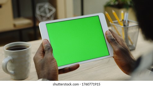 Over shoulder view on the white tablet computer holded by african american man horizontally. Fingers scrolling and taping on it. Green screen. Chroma key. Wooden office desk with cup of coffee