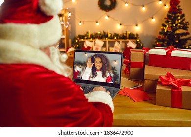 Over Shoulder View Santa Claus Or Father Christmas Having Online Video Call With Happy African American Girl On Laptop Computer In His Workshop. Self-isolation And Virtual Celebration At Home Concept