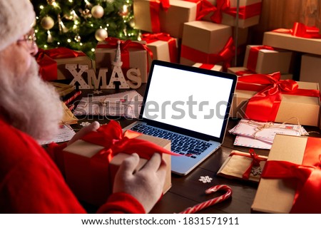 Over shoulder view of Santa Claus holding xmas gift using laptop computer with white blank empty mock up screen monitor for e commerce website ad sitting at decorated table on Merry Christmas eve.