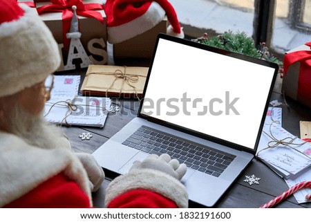Over shoulder view of Santa Claus wearing costume using laptop computer with white blank empty mock up screen monitor sitting at workshop table on Merry Christmas eve. E commerce website ads concept