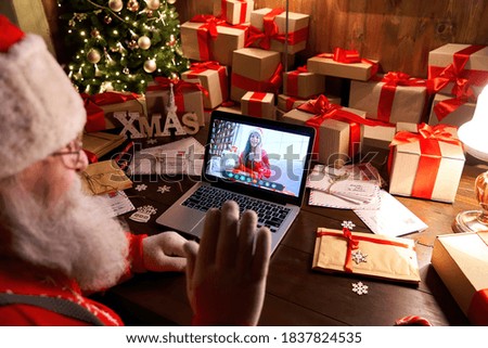 Over shoulder view of Santa Claus video calling kid girl on laptop greeting child by webcam talk open Christmas gift box by virtual online videocall using computer sit at workshop table on xmas eve.