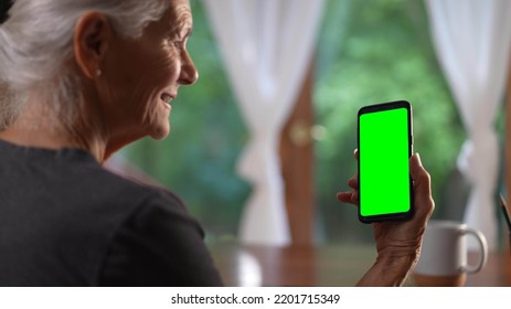 Over Shoulder View Of Smiling Senior Elderly Woman Making Video Call On Smartphone Phone With Green Screen Talking And Listening During Video Chat With Friends Or Colleagues.