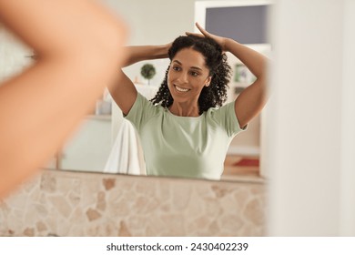 Over the shoulder view of a smiling young African woman tying her long curly hair back in a bathroom mirror at home in the morning - Powered by Shutterstock