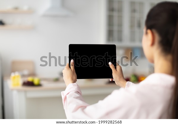 Over The Shoulder View Of Unrecognizable Woman
Using Digital Tablet With Empty Screen Standing In Kitchen At Home.
Application Or Internet Website Advertising, Mock Up, Free Space,
Selective Focus