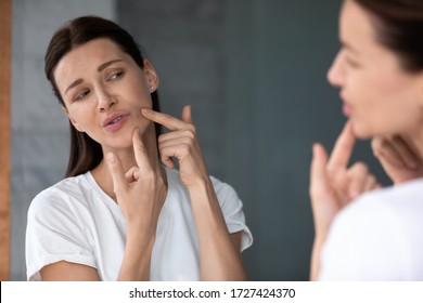 Over shoulder view upset female face reflected in mirror, woman touch face squeezes pimple caused by hormonal imbalance changes, stress or diet. Skin problem, jawline acne or new mole appears concept