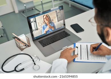 Over Shoulder View Of Videocall Chat With Female Older Patient And Indian Male Doctor In Modern Clinic Hospital Using Laptop Computer, Consulting Online Remotely. Telemedicine Healthcare Concept.