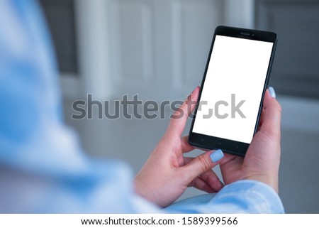 Over shoulder view: woman hands holding black smartphone with white blank screen in home interior. Mock up, copyspace, template, entertainment and technology concept
