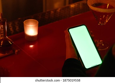 Over The Shoulder View Of Woman Holding Green Screen Phone At Bar At Night.