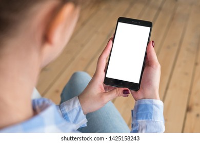Over shoulder view: woman in plaid shirt holding black smartphone with white blank screen in home interior with floorboards. Mock up, copyspace, template, entertainment and technology concept